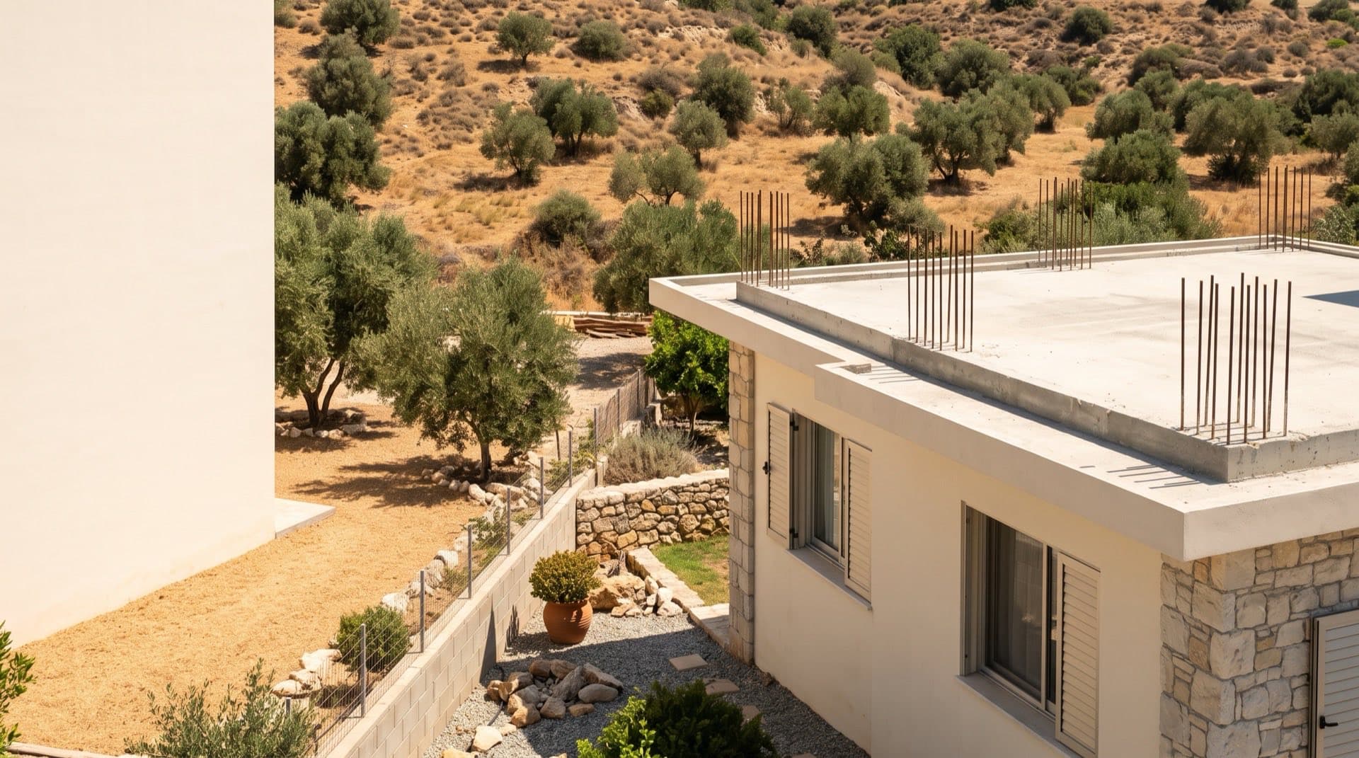 A single-storey rendered Cyprus house with exposed vertical rebar starter bars rising from the roof slab edge, olive hillside behind, in bright midday sun.